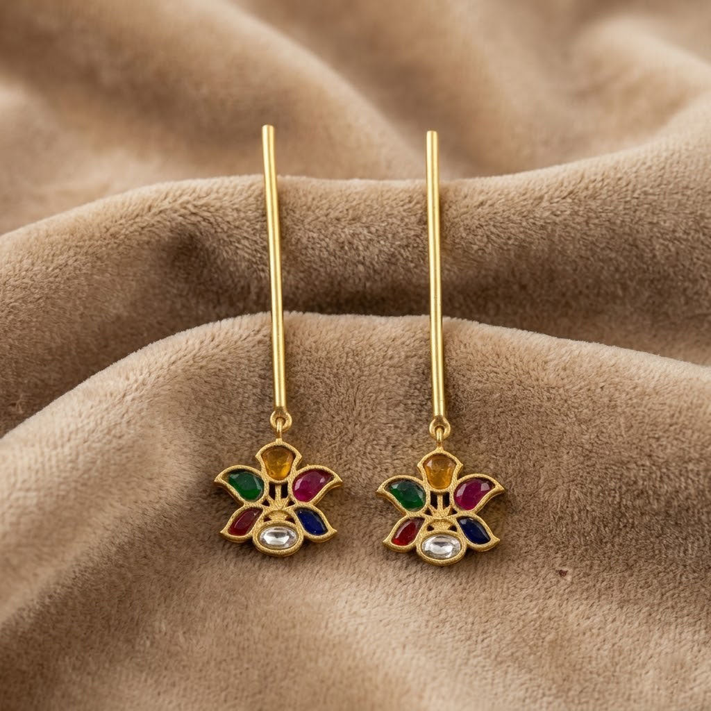 A pair of gold-tone dangle earrings with a small flower-shaped charm featuring multicolor enamel details and a clear central stone, displayed on a textured brown fabric background.