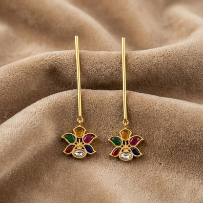 A pair of gold-tone dangle earrings with a small flower-shaped charm featuring multicolor enamel details and a clear central stone, displayed on a textured brown fabric background.
