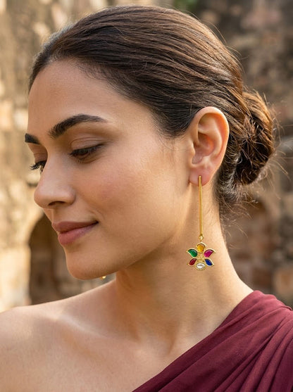 A woman in profile wearing a gold dangle earring with a colorful flower-like charm, set against a blurred stone wall background.