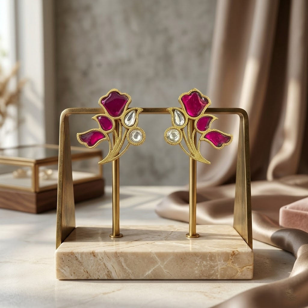 A pair of gold-tone floral stud earrings with deep pink enamel petals and clear teardrop crystals, displayed on a gold stand with a marble base in a softly lit interior setting.