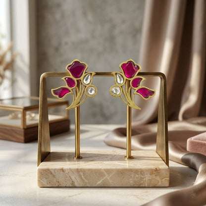 A pair of gold-tone floral stud earrings with deep pink enamel petals and clear teardrop crystals, displayed on a gold stand with a marble base in a softly lit interior setting.