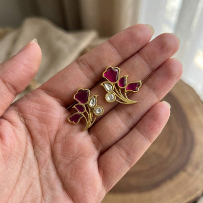 A pair of gold floral stud earrings with deep pink enamel petals and small clear stones, held in a person's hand.