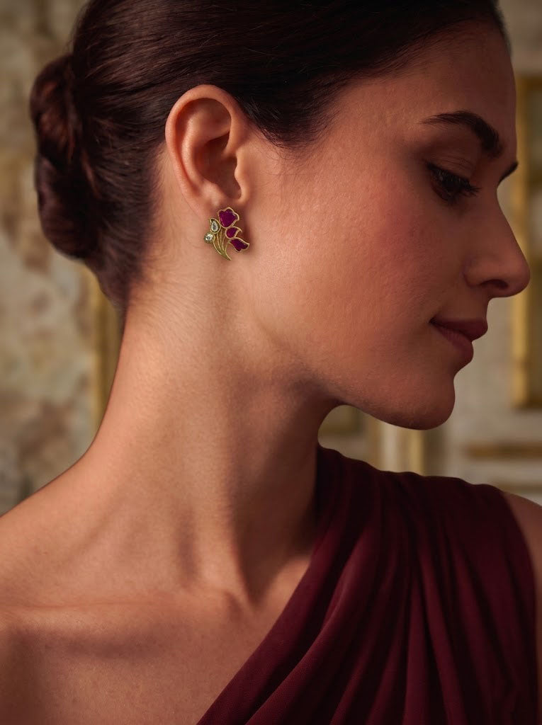 A close-up side profile of a woman wearing a small butterfly-shaped earring with pink and green enamel details, paired with a draped burgundy top.