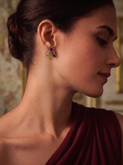 A close-up side profile of a woman wearing a small butterfly-shaped earring with pink and green enamel details, paired with a draped burgundy top.