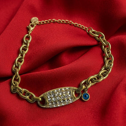 A gold chain bracelet with a central oval plate paved with clear crystals and a small blue evil eye charm, displayed on a red satin fabric background.